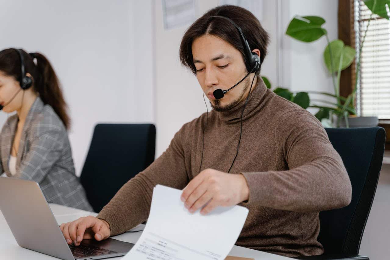 Operatore di telemarketing B2B con cuffie e microfono al lavoro su laptop e documenti in ufficio moderno.